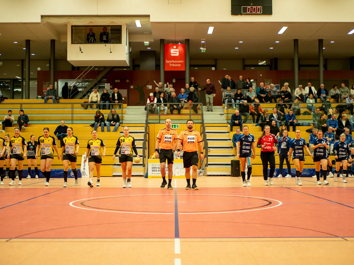 Beide Teams von links nach rechts aufgereiht, dazwischen die Schiedsrichter. Die Reihe läuft von der Tribüne weg auf die Kamera zu aufs Spielfeld.
