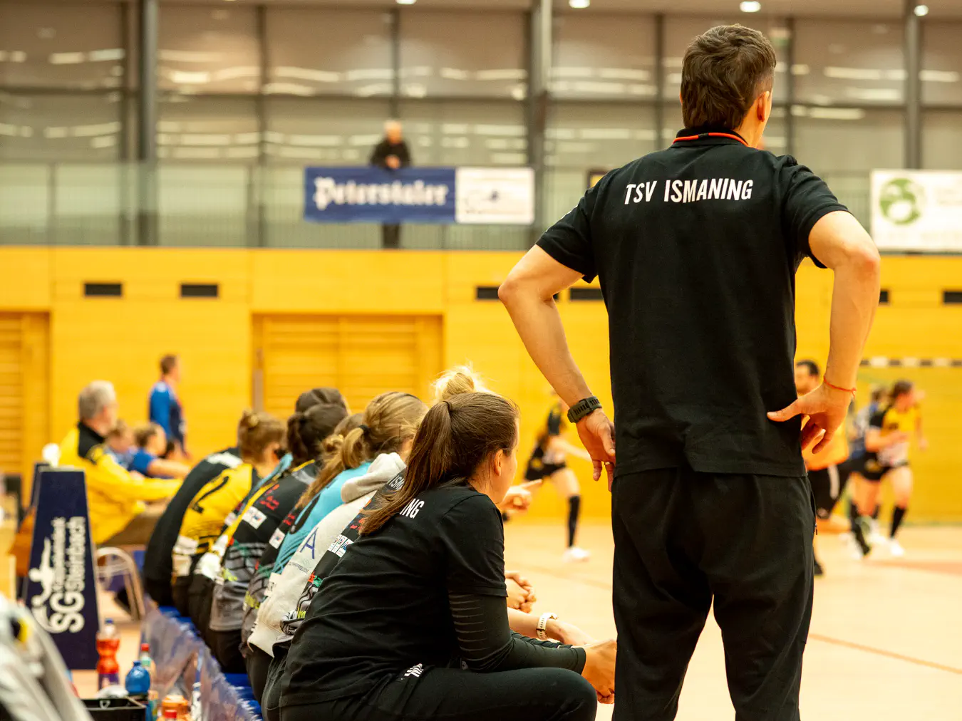 Eine Reihe von Menschen von hinten. Links sitzen einige Menschen auf einer Bank, rechts neben der Bank steht ein Mensch. Im Hintergrund Spielgeschehen.