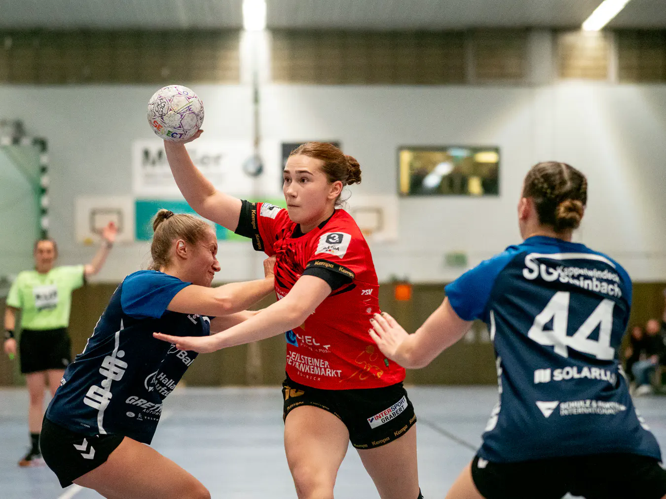 Mittig eine Schozacher Spielerin im Angriff, den Arm zum Wurf ausholend. Links und rechts zwei Kappelwindecker Verteidigerinnen.