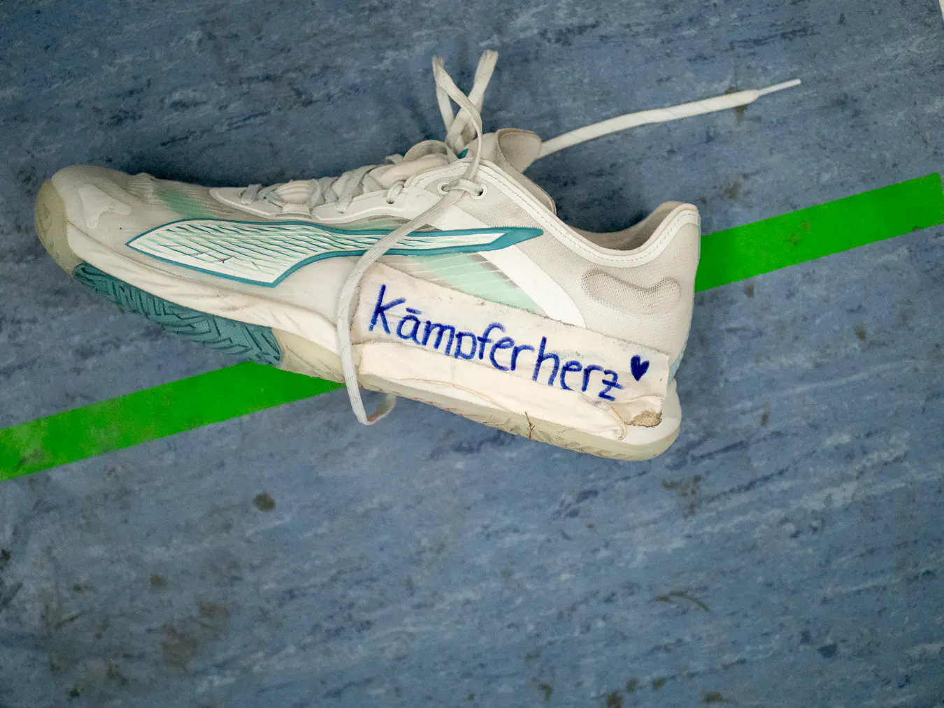 Ein einzelner Sportschuh liegt auf dem Hallenboden. Auf die Seite ist Tape geklebt, darauf steht 'Kämpferherz' geschrieben.