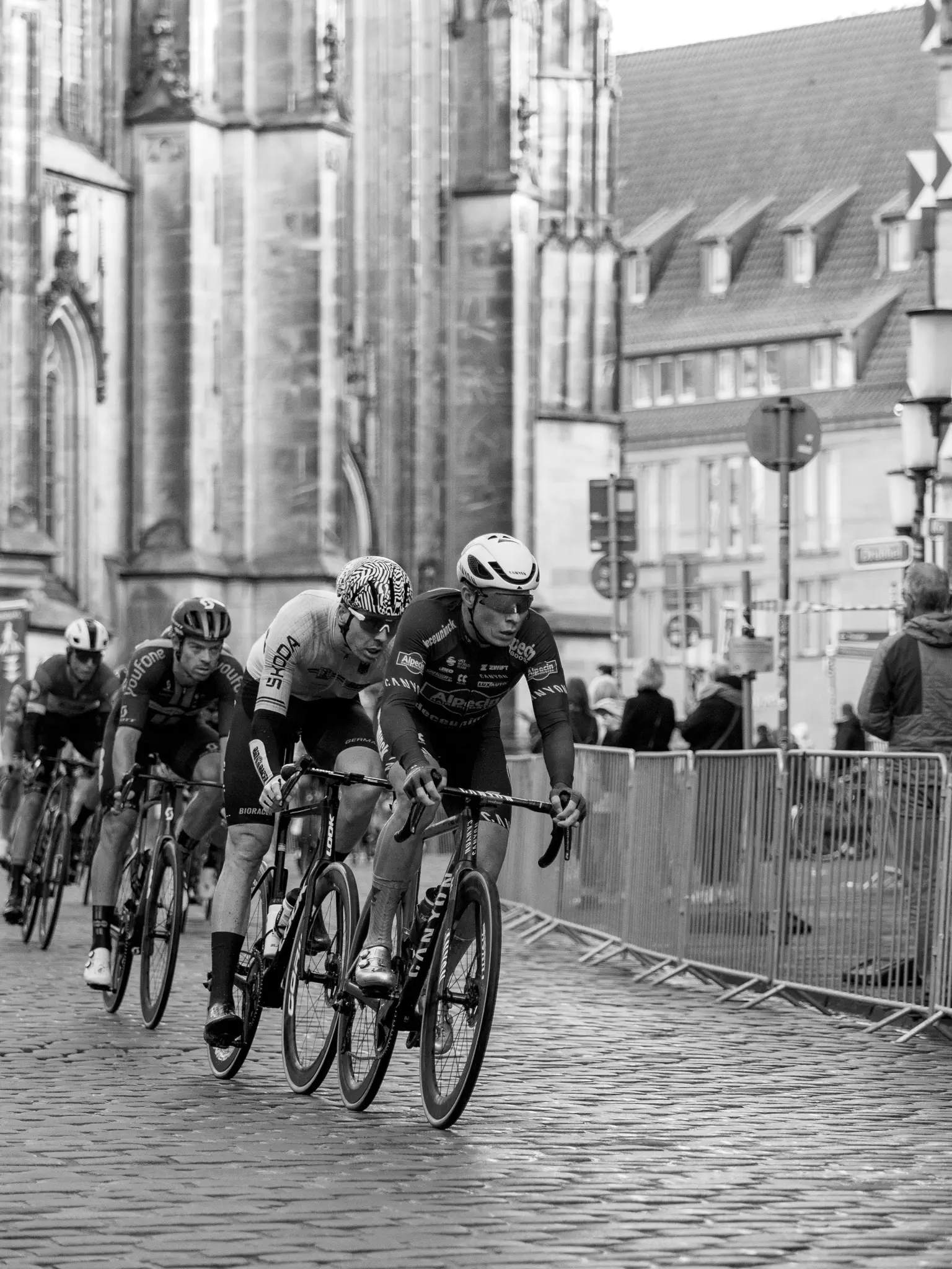 Eine Gruppe Radfahrer kommt dem Betrachter entgegen, im Hintergrund ein aufwändiges Backsteingebäude