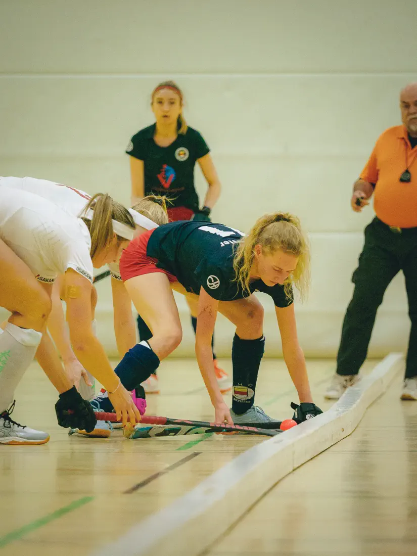 Eine Frau im schwarzen Trikot, gebückt, hält ihren Hockeyschläger flach über den Boden; eine Frau im weißen trikot versucht, einen Ball daran vorbei zu spielen