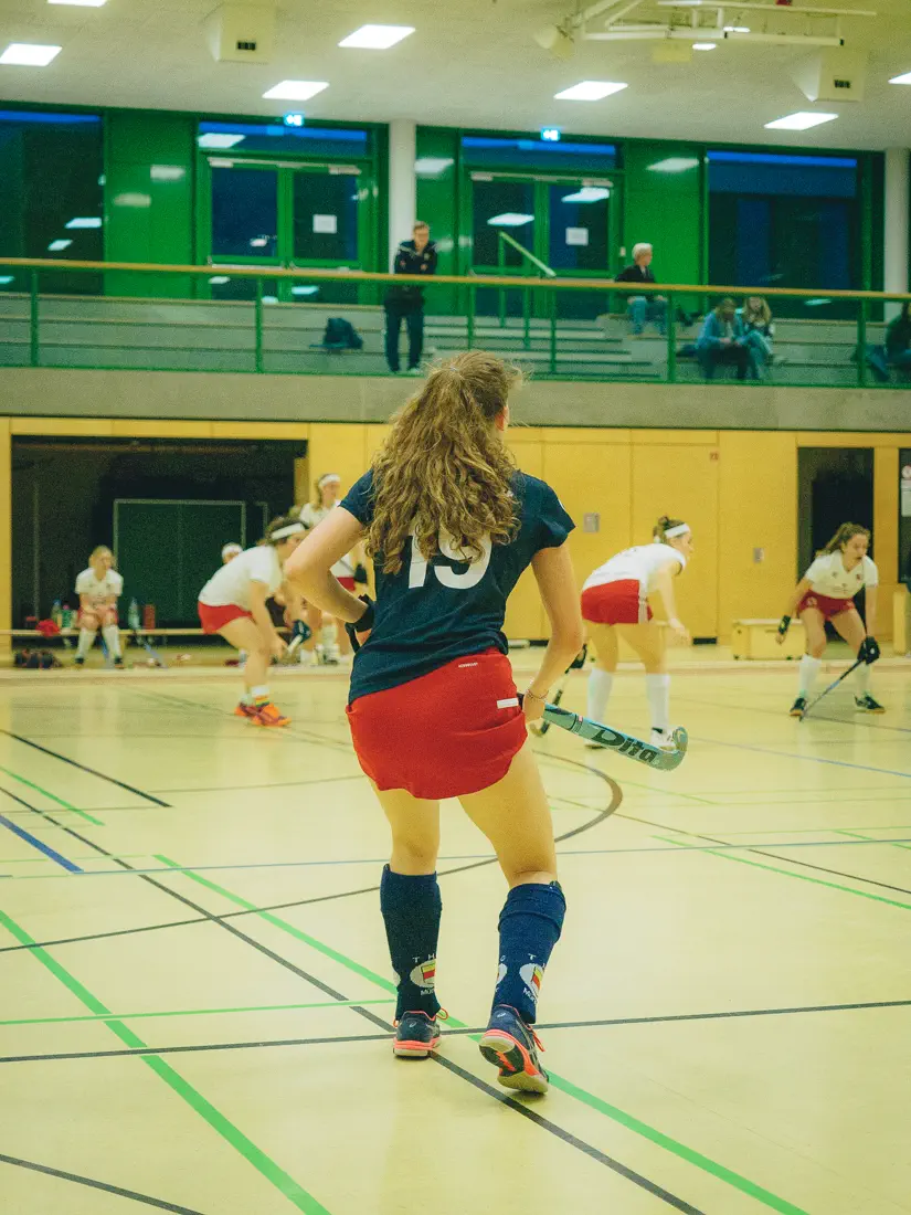 Eine Frau im schwarzen Trikot von hinten, dahinter das Spielfeld und drei Frauen in weißen Trikots