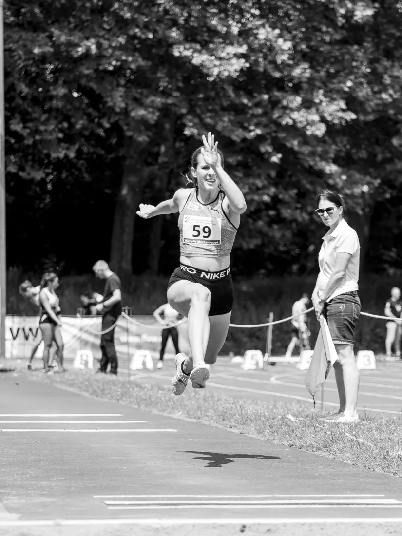 Eine Frau im Sprung von schräg vorn, nach schräg links die Anlaufbahn, rechts eine Kampfrichterin beobachtend