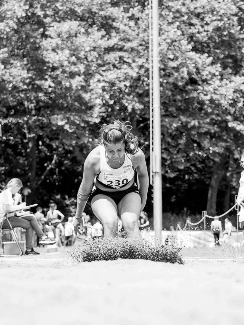 Eine Frau von vorn bei der Landung in einer Sandgrube, die Füße gerade aufgekommen, Sand spritz hoch und verdeckt Füße und einen Teil der Unterschenkel, der Oberkörper nach vorn gebeugt, im Hintergrund eine Kampfrichterin und Bäume