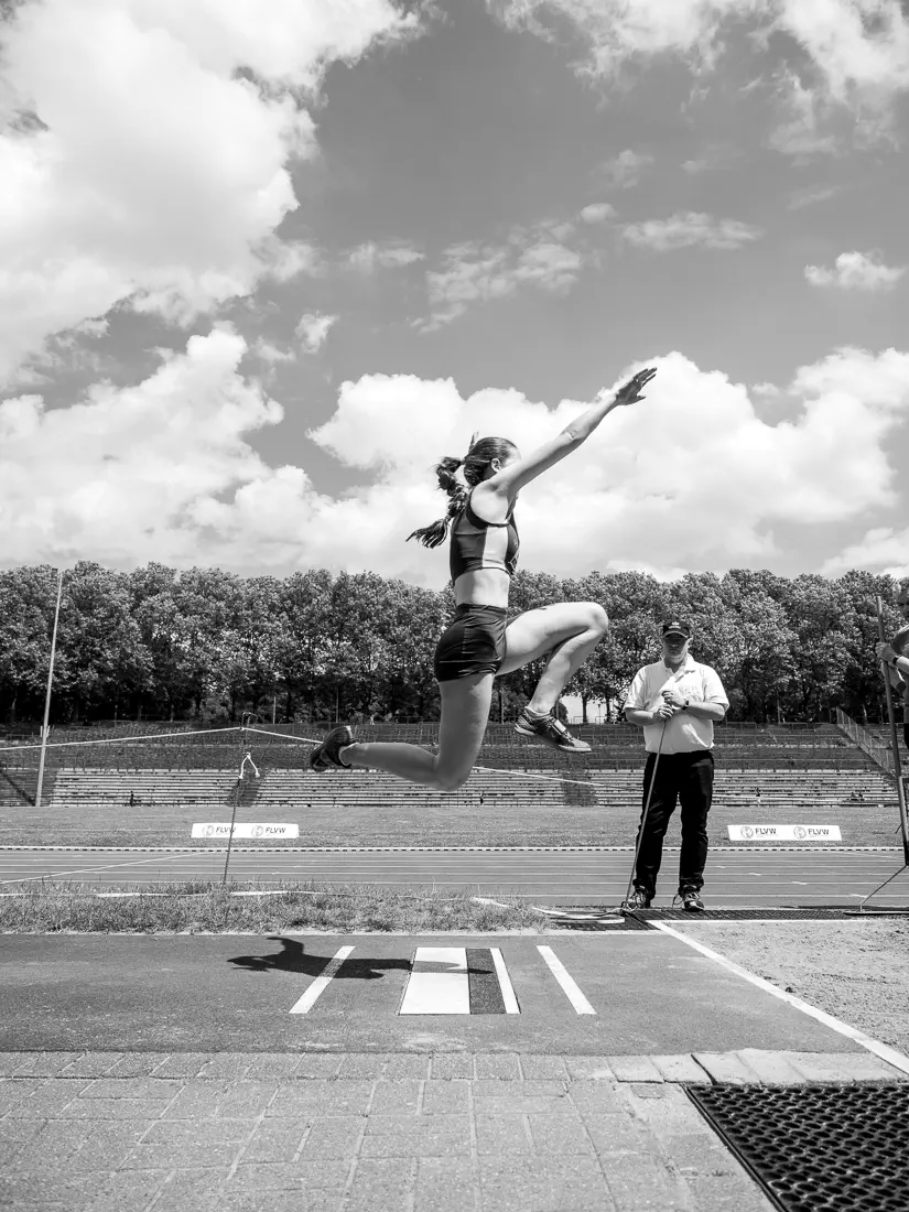 Eine Frau im Sprung von der Seite, die Arme nach schräg vorn gestreckt, die Füße angezogen, unter ihr der Absprugbalken, im Hintergrund eine Tribüne, Bäume und bewölkter Himmel
