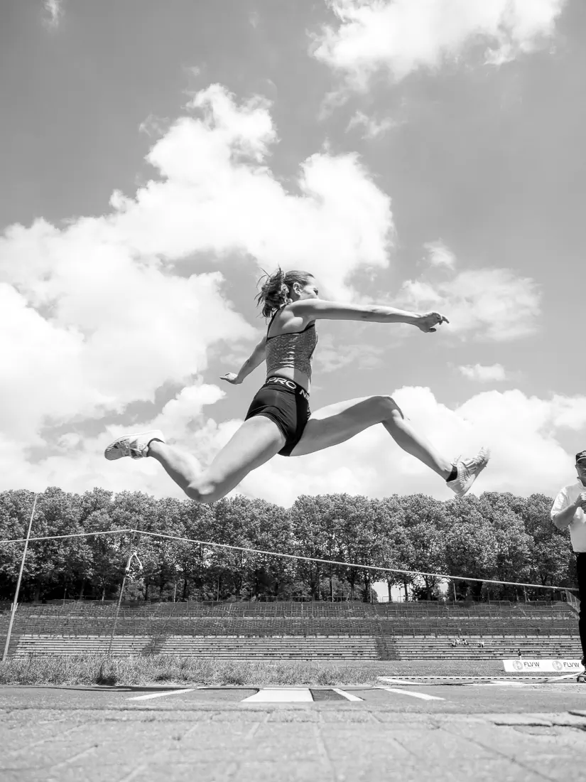 Eine Frau im Sprung von der Seite, unter ihr der Absprungbalken, im Hintergrund eine Tribüne, Bäume und bewölkter Himmel