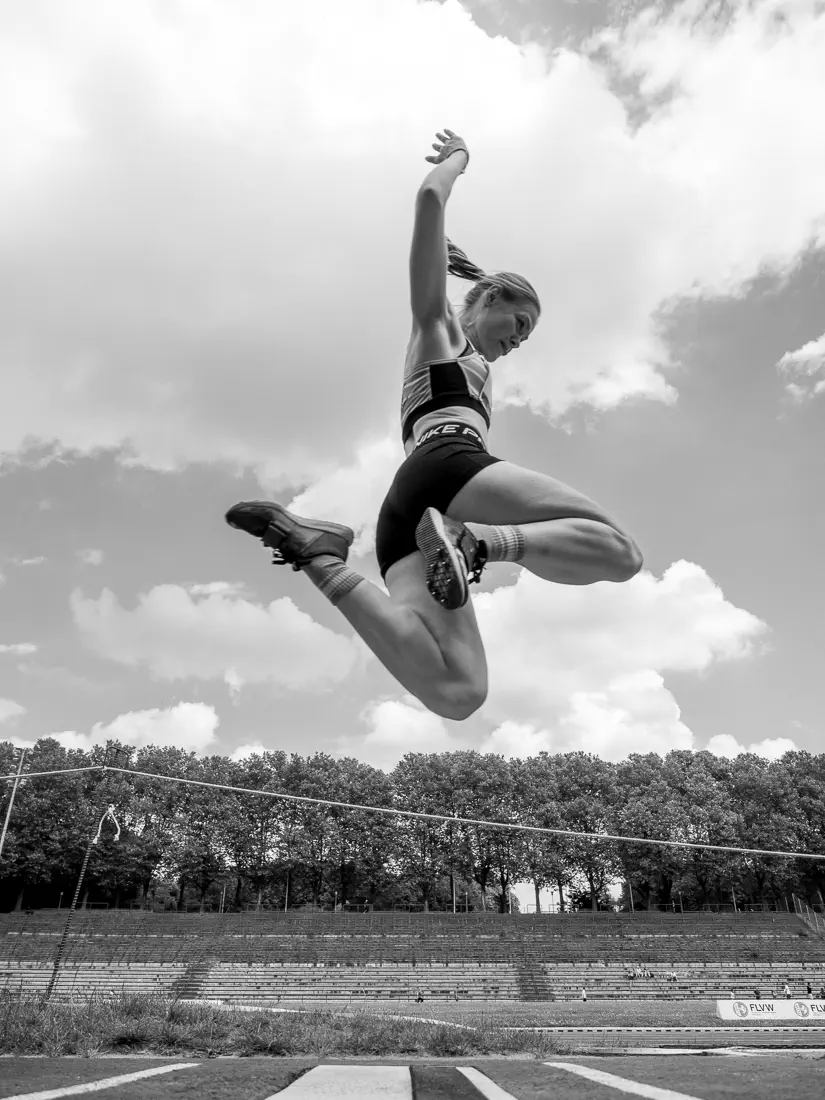 Eine Frau im Sprung von der Seite, die Arme nach oben gestreckt, die Füße angezogen, unter ihr der Absprugbalken, im Hintergrund eine Tribüne, Bäume und bewölkter Himmel