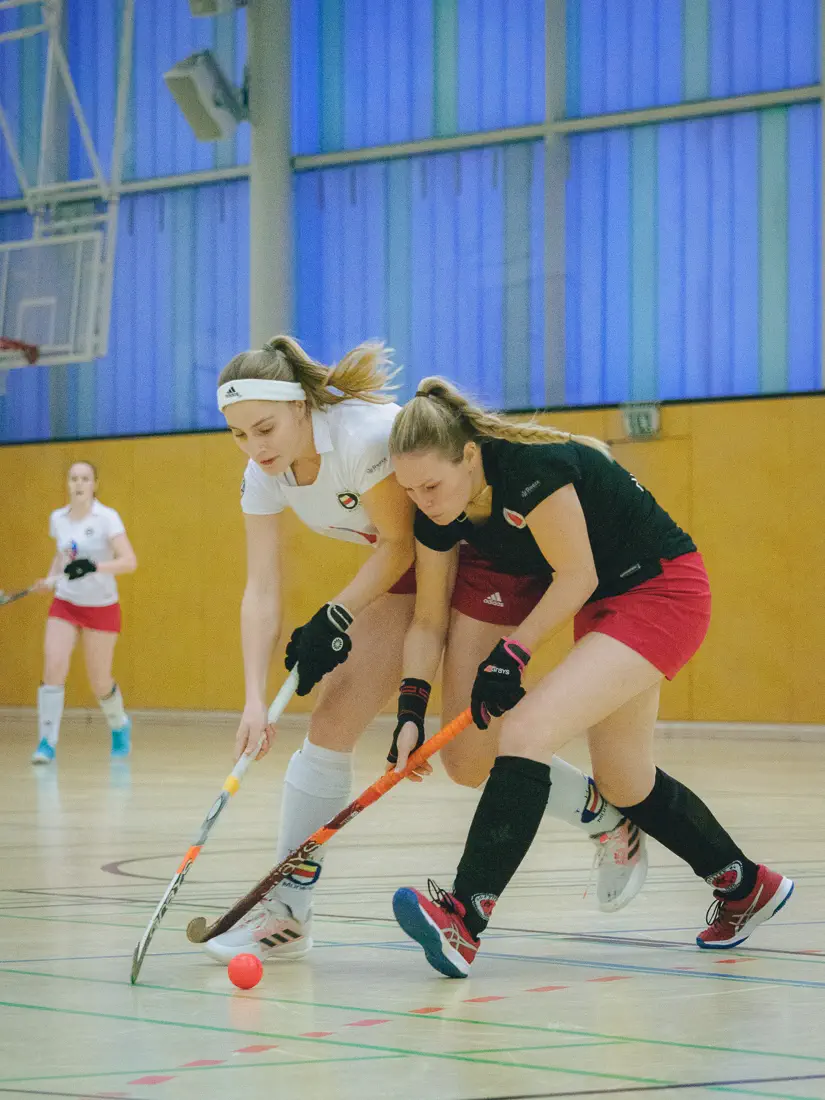 Eine Frau im schwarzen Trikot von links vorn, nach vorn gebeugt, treibt mit einem Hockeyschläger einen Hockeyball; eine Frau im weißen Trikot links daneben versucht, den Ball mit ihrem Schläger zu erreichen