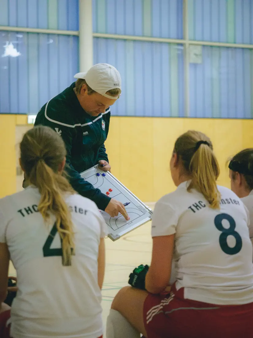Ein Mann von rechts vorn, hält eine Magnettafel mit einem aufgemalten Hockeyfeld, zeigt darauf; davor drei Frauen von hinten, die auf die Tafel schauen