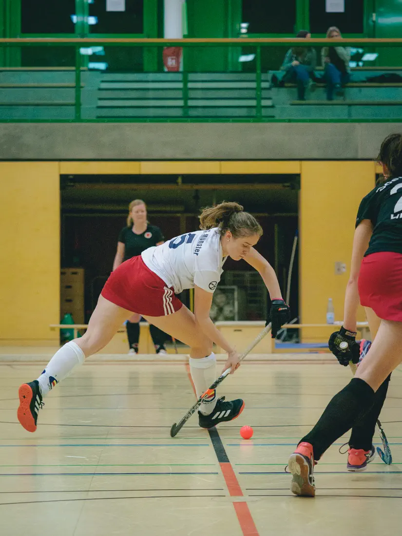 Eine Frau im weißen Trikot, von links nach rechts durchs Bild laufend, hat mit dem Schläger ausgeholt und ist kurz davor, einen Ball nach rechts aus dem Bild zu spielen
