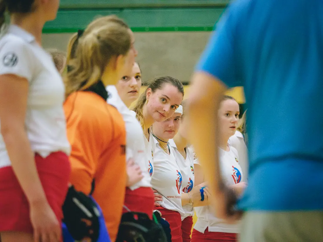 Eine Reihe von Frauen in weißen Trikots, alle schauen nach rechts, eine beugt sich vor und schaut die Reihe entlang dem Betrachter entgegen