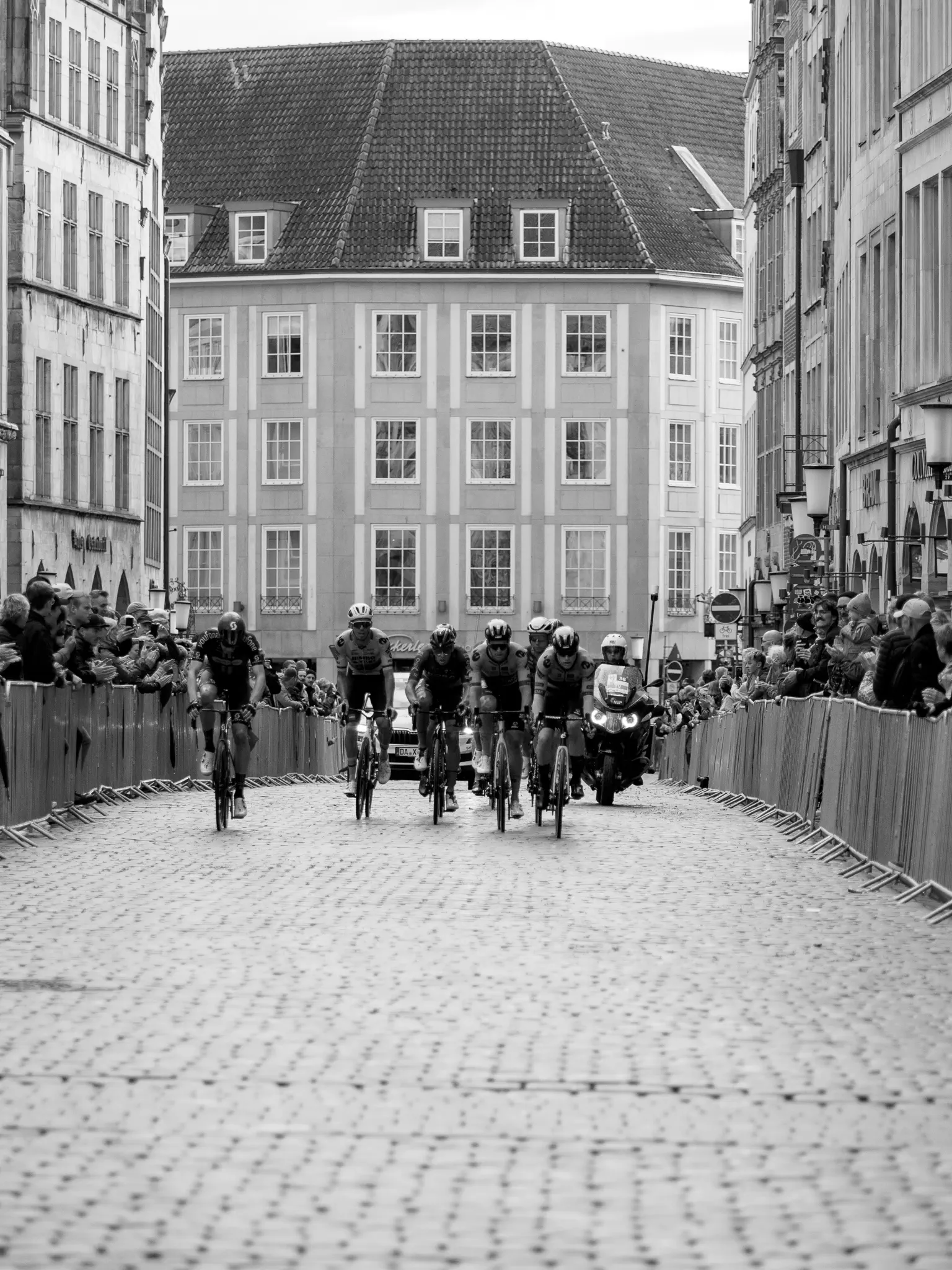 Eine Häuserschlucht, darin mehrere Radfahrer nebeneinander, die auf den Betrachter zu fahren