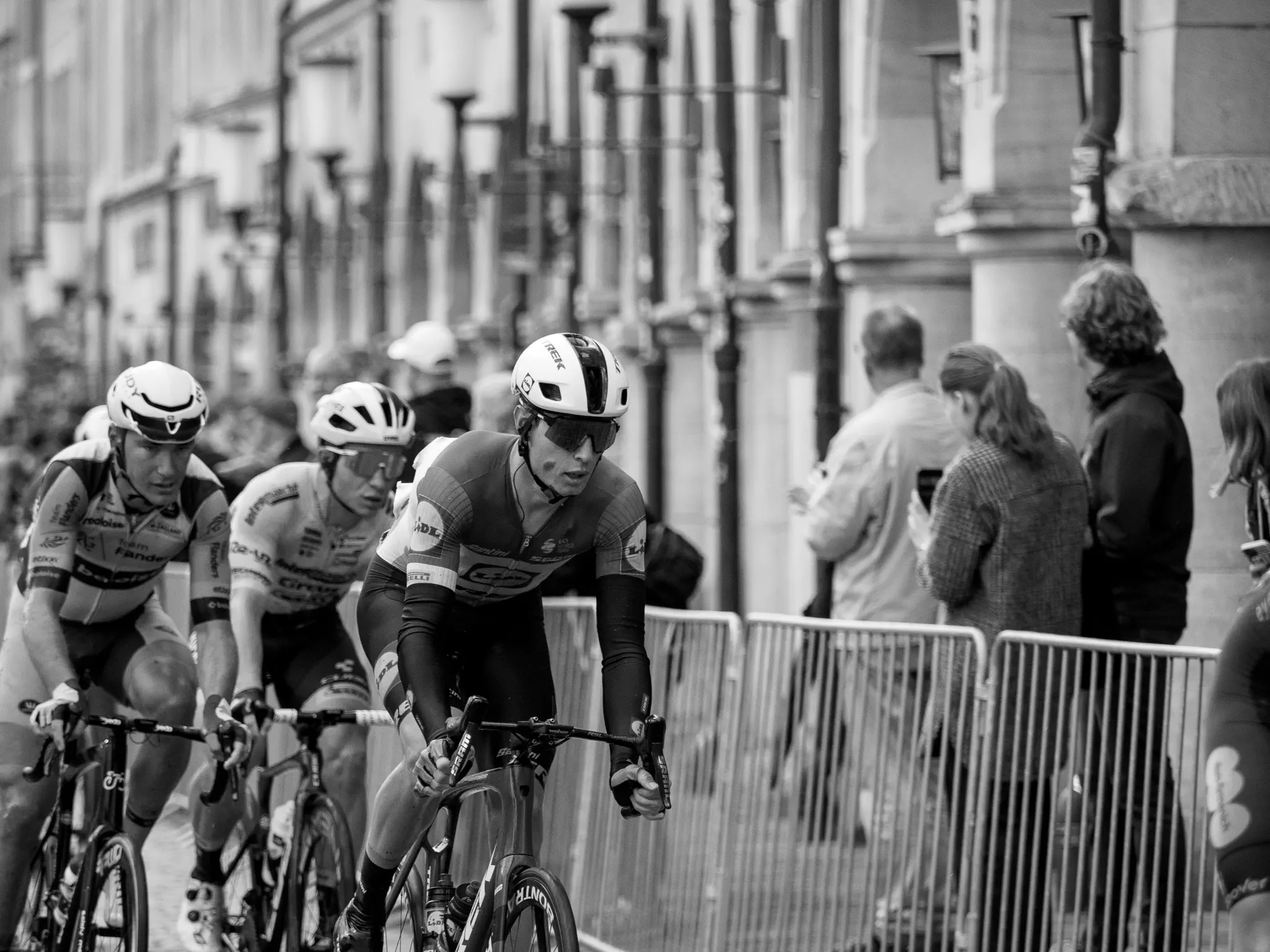 Drei Radfahrer kommen von schräg links hinten in Richtung Bildmitte dem Betrachter entgegen, dahinter Absperrgitter, dahinter Zuschauer