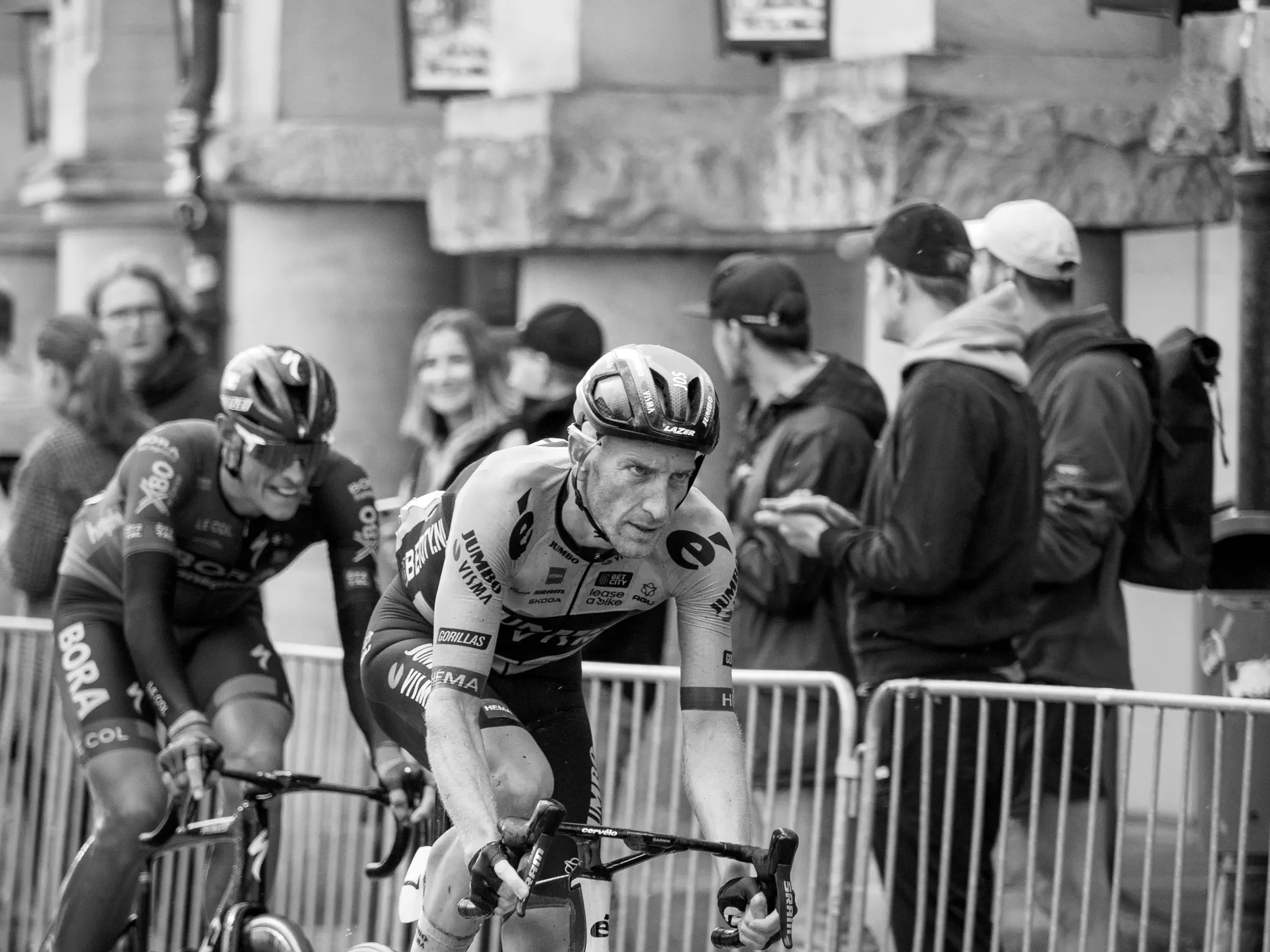 Zwei Radfahrer kommen von schräg links hinten in Richtung Bildmitte dem Betrachter entgegen, dahinter Absperrgitter, dahinter Zuschauer