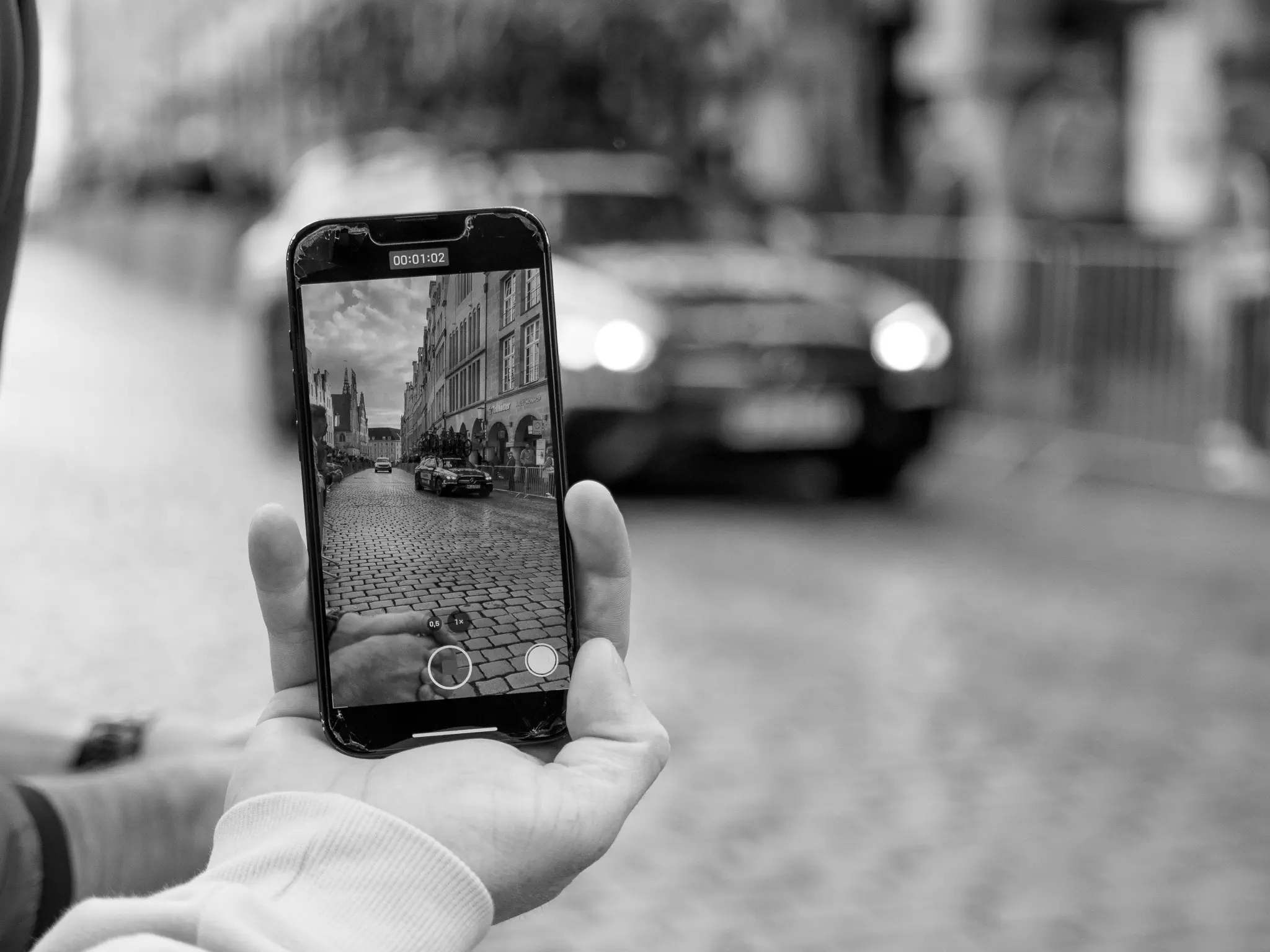 Ein Smartphone, darauf geöffnet die Kamera-App mit Blick auf Autos in einer Häuserschlucht; dahinter unscharf ein Auto in derselben Häuserschlucht