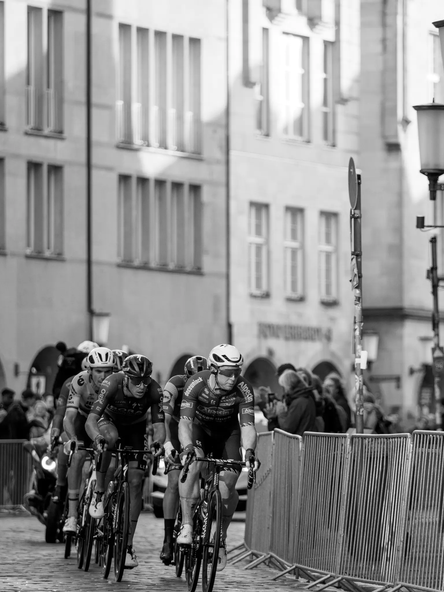 Eine Gruppe Radfahrer kommt dem Betrachter entgegen, seitlich rechts Absperrzäune