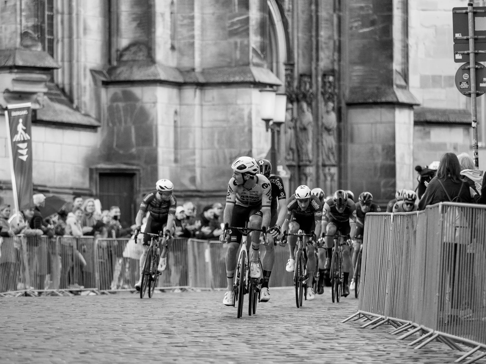 Eine Gruppe Radfahrer kommt dem Betrachter entgegen, im Hintergrund ein aufwändiges Backsteingebäude