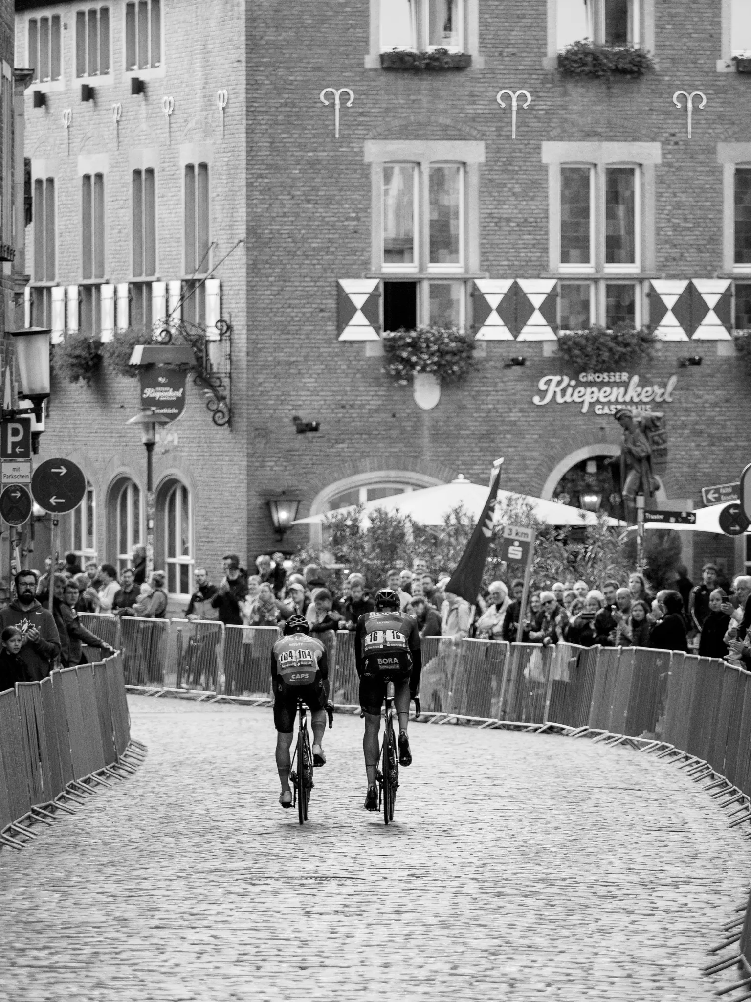 Zwei Radfahrer von hinten fahren durch eine Gasse von Absperrgittern, beidseitig Zuschauer