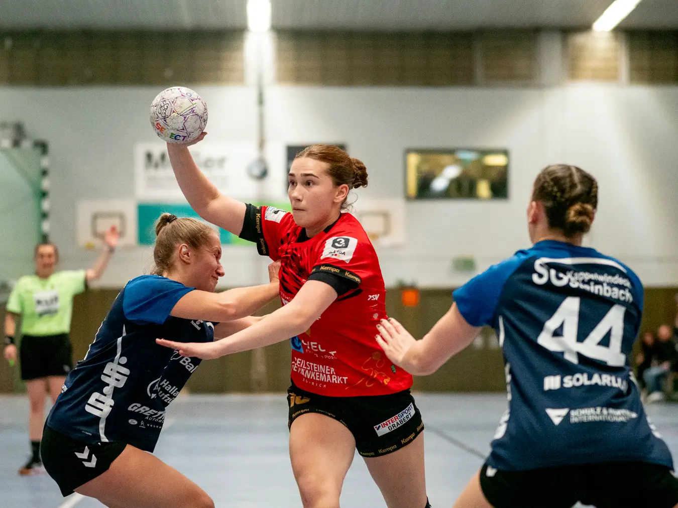 Mittig eine Schozacher Spielerin im Angriff, den Arm zum Wurf ausholend. Links und rechts zwei Kappelwindecker Verteidigerinnen.