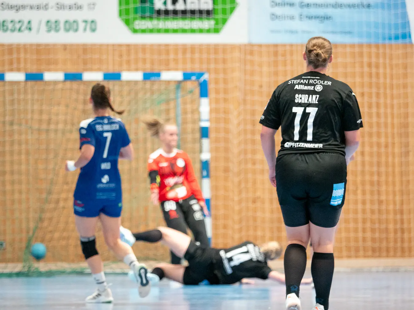 Im Hintergrund ein Handball im Tornetz, davor die Torfrau in der Reaktion, davor eine Angreiferin auf dem Boden liegend, davor eine Angreiferin zusehend