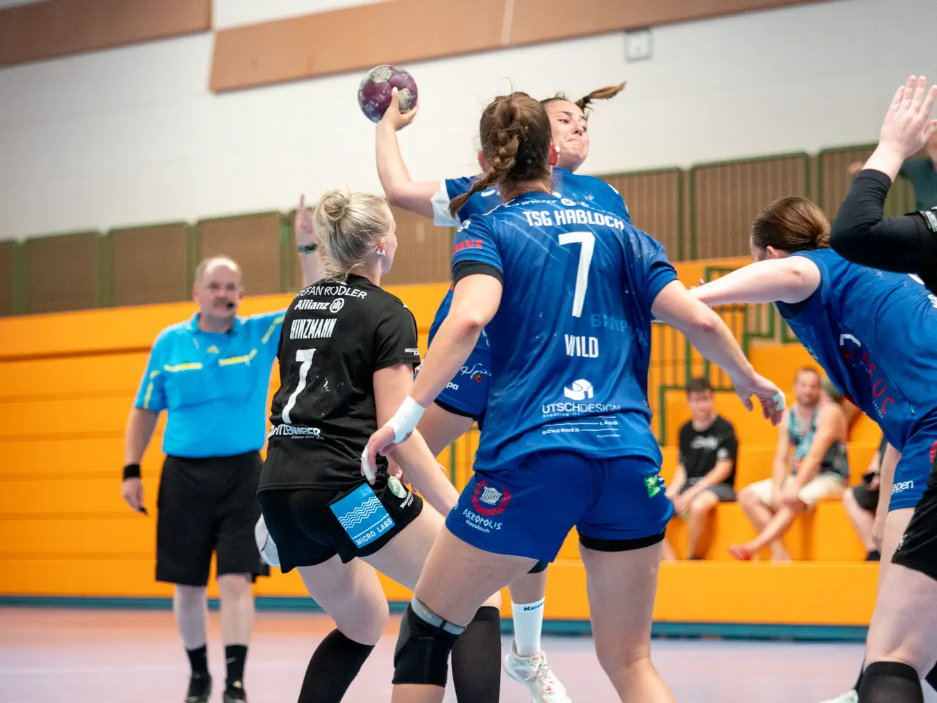 Zwei Frauen von hinten, dahinter eine Frau im Sprungwurf, dahinter ein Schiedsrichter mit einem erhobenen Finger