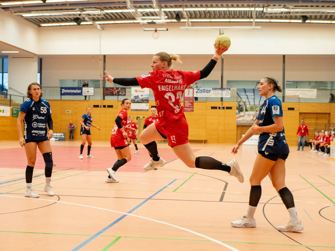 Eine Frau im Sprungwurf von rechts nach links, zwei Frauen schauen von links und rechts zu.