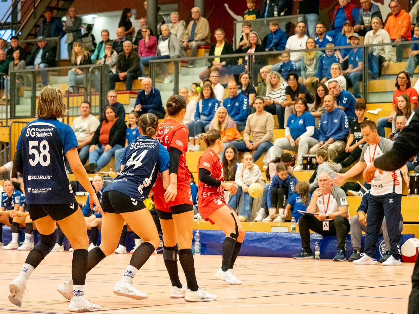 Einige Frauen von hinten schauen einem Ball nach, der in Richtung Seitenlinie fliegt. Im Hintergrund Publikum auf einer Tribüne.