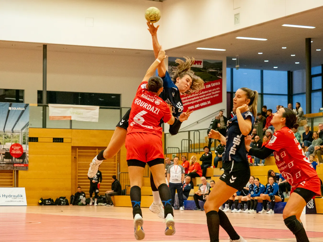 Eine Frau von vorn im Sprungwurf, davor eine Frau von hinten, ebenfalls springend, in der Verteidigung.