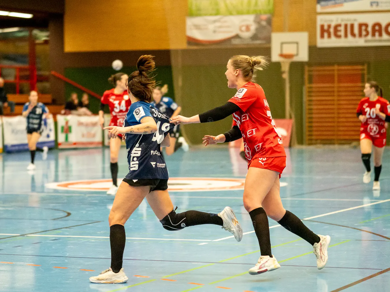 Eine Spielerin im blauen Trikot läuft nach links durchs Bild. Eine Spielerin im roten Trikot folgt ihr am Trikot ziehend.