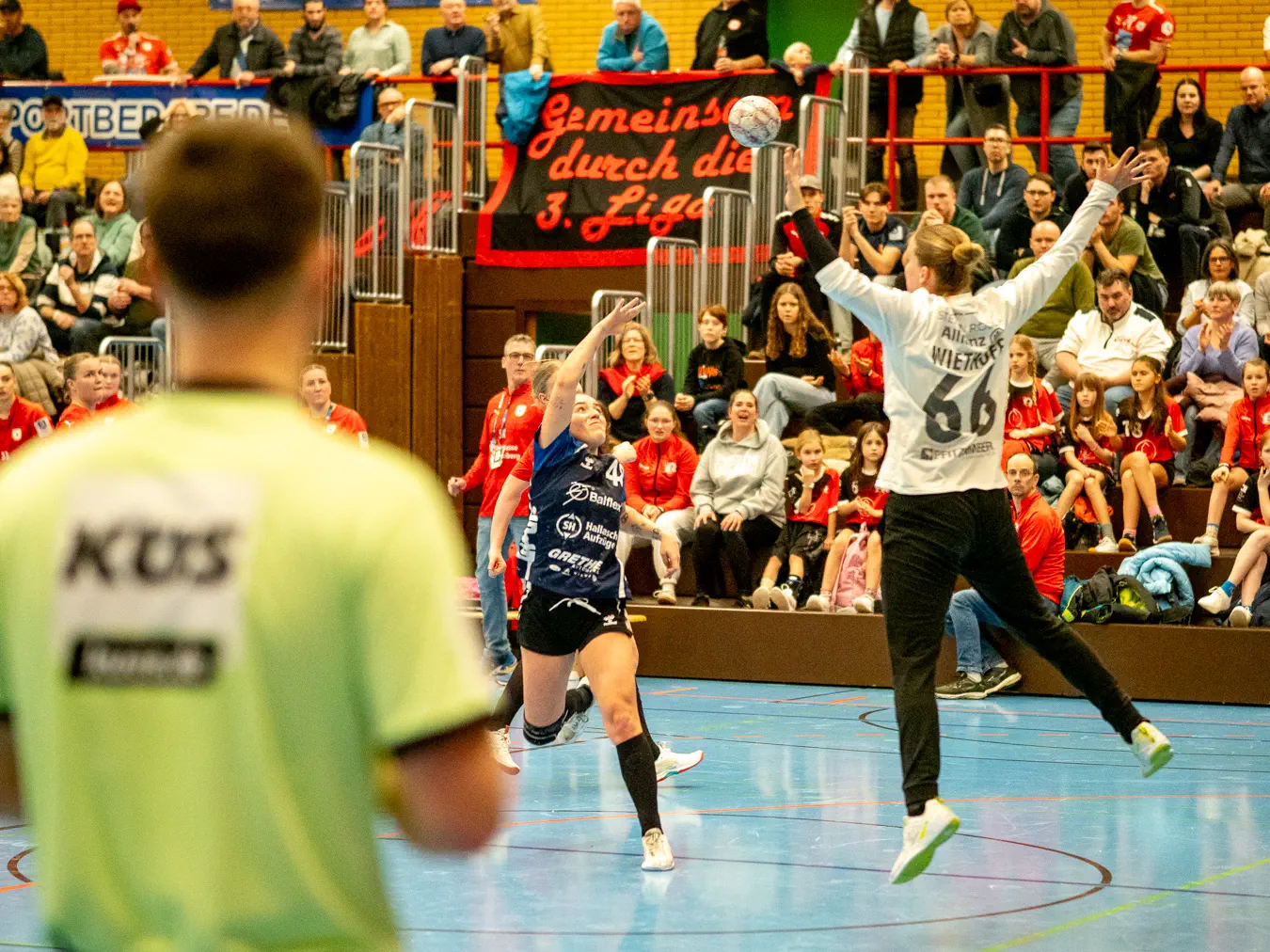 Eine Spielerin im blauen Trikot wirft einen Heber über eine springende Torfrau im weißen Trikot.