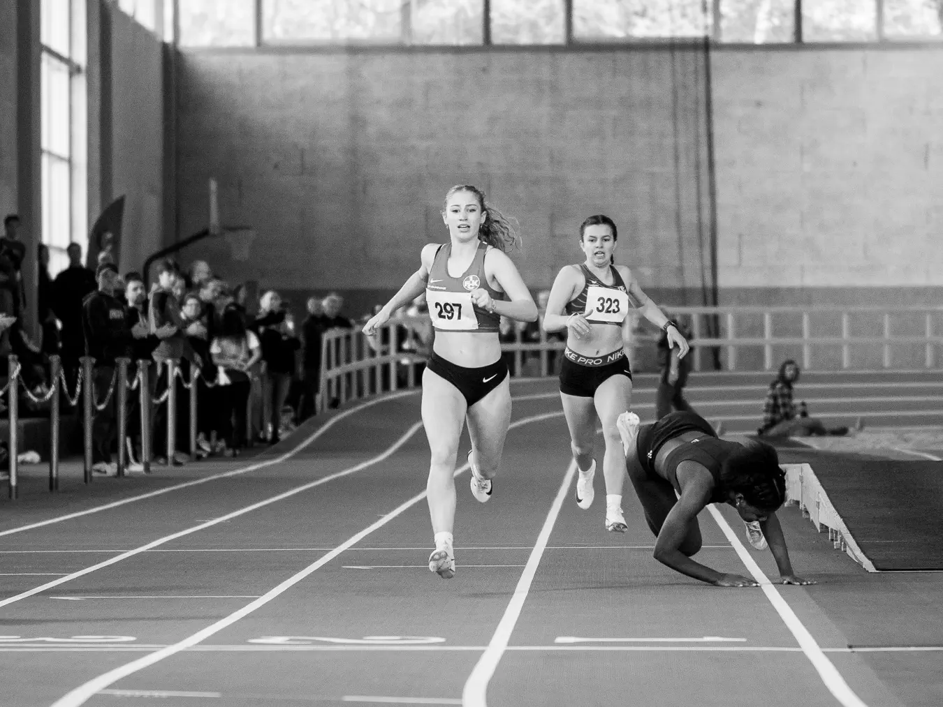 Zwei Athletinnen auf einer Rundbahn, von vorn, kurz vor dem Zieleinlauf. Rechts daneben eine weitere Athletin mitten im Sturz, die Arme zum Abfangen ausgestreckt.