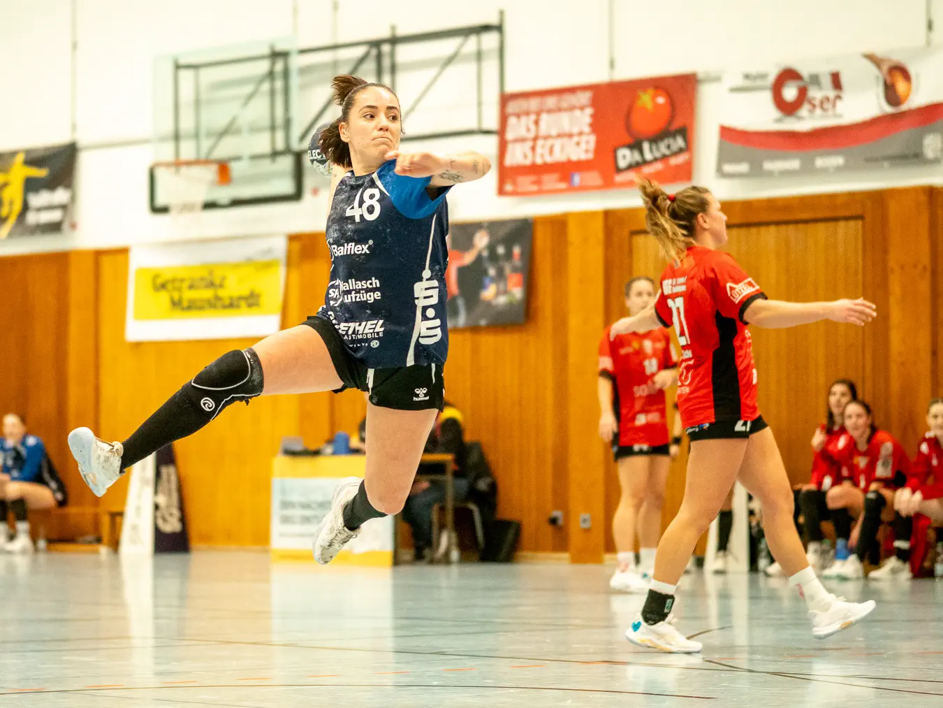 Eine Spielerin im blauen Trikot im Sprungwurf, mit einigem Abstand im Hintergrund Spielerinnen in roten Trikots.