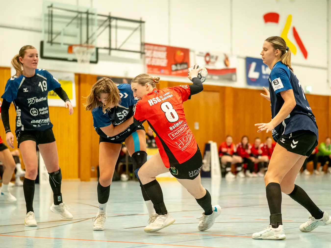 Eine Spielerin im roten Trikot holt zum Wurf aus, eine Spielerin im blauen Trikot versucht sie von der Seite festzuhalten.