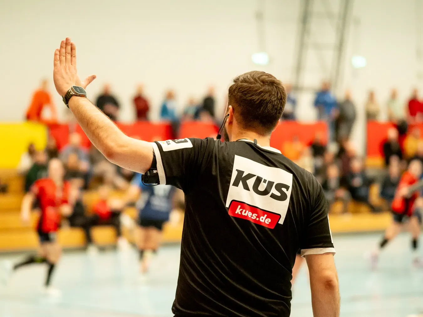 Ein Schiedsrichter im schwarzen Trikot hebt die linke flache Hand. Im Hintergrund unscharf Spielgeschehen.