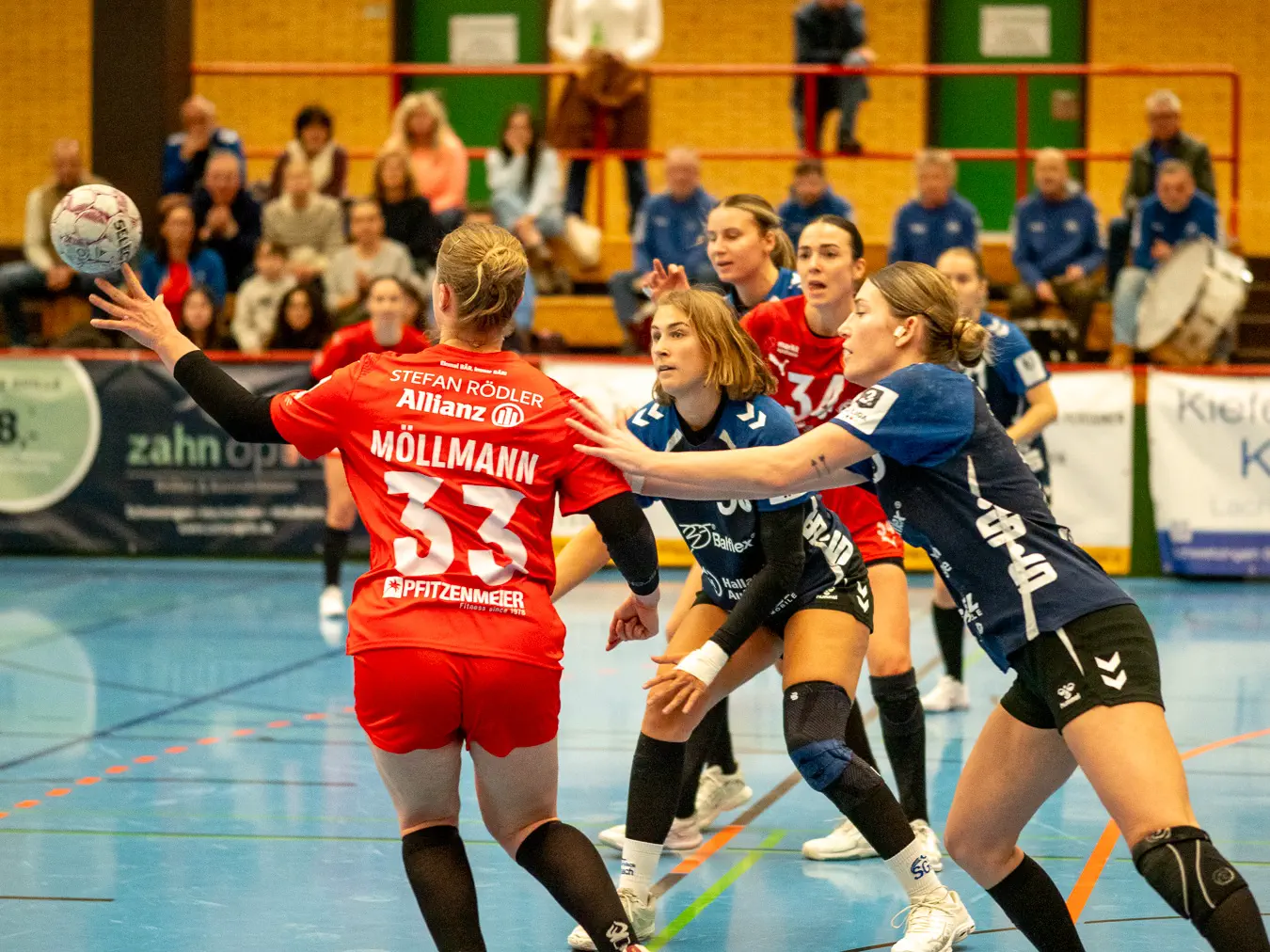 Eine Spielerin im roten Trikot passt den Ball und wird dabei von zwei Spielerinnen in blauen Trikots bedrängt.