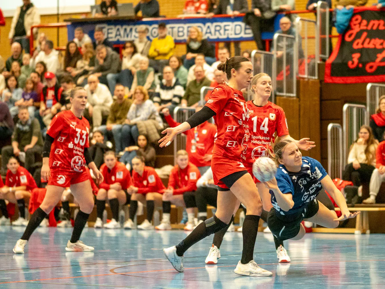 Eine Spielerin im blauen Trikot im Sprungwurf kurz vor dem Boden. Zwei Spielerinnen in roten Trikots sehen zu, strecken die Arme seitlich von sich.