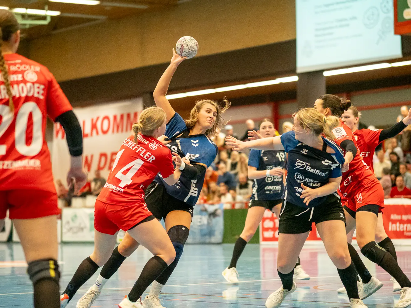 Zwei Spielerinnen in blauen Trikots beim Passversuch werden von zwei Spielerinnen in roten Trikots bedrängt.