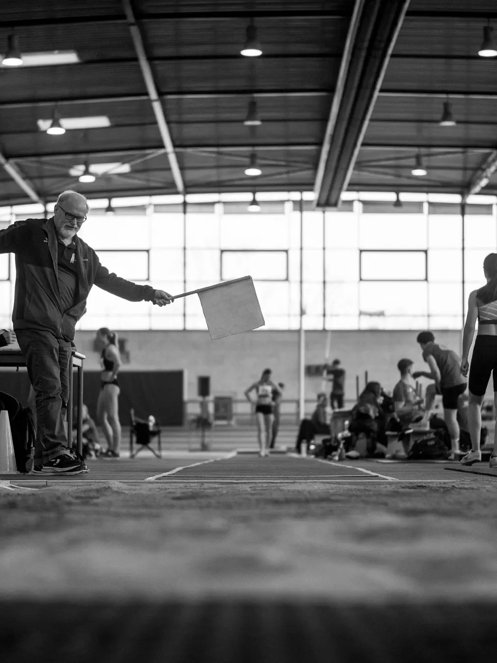 Ein älterer Mann hält eine helle Fahne über eine Laufbahn vor einer Sandgrube. Im Hintergrund wartet eine Athletin auf ihren Anlauf.