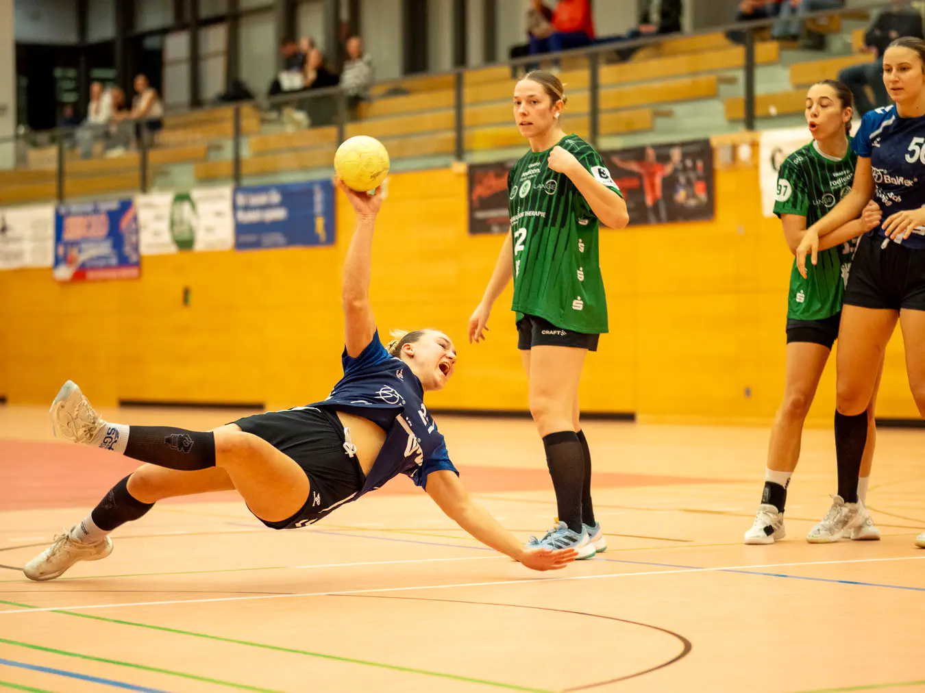 Eine Angreiferin mit Ball in der Hand im Fallen, der Körper waagerecht zum Boden in der Luft, drei andere Spielerinnen schauen zu.
