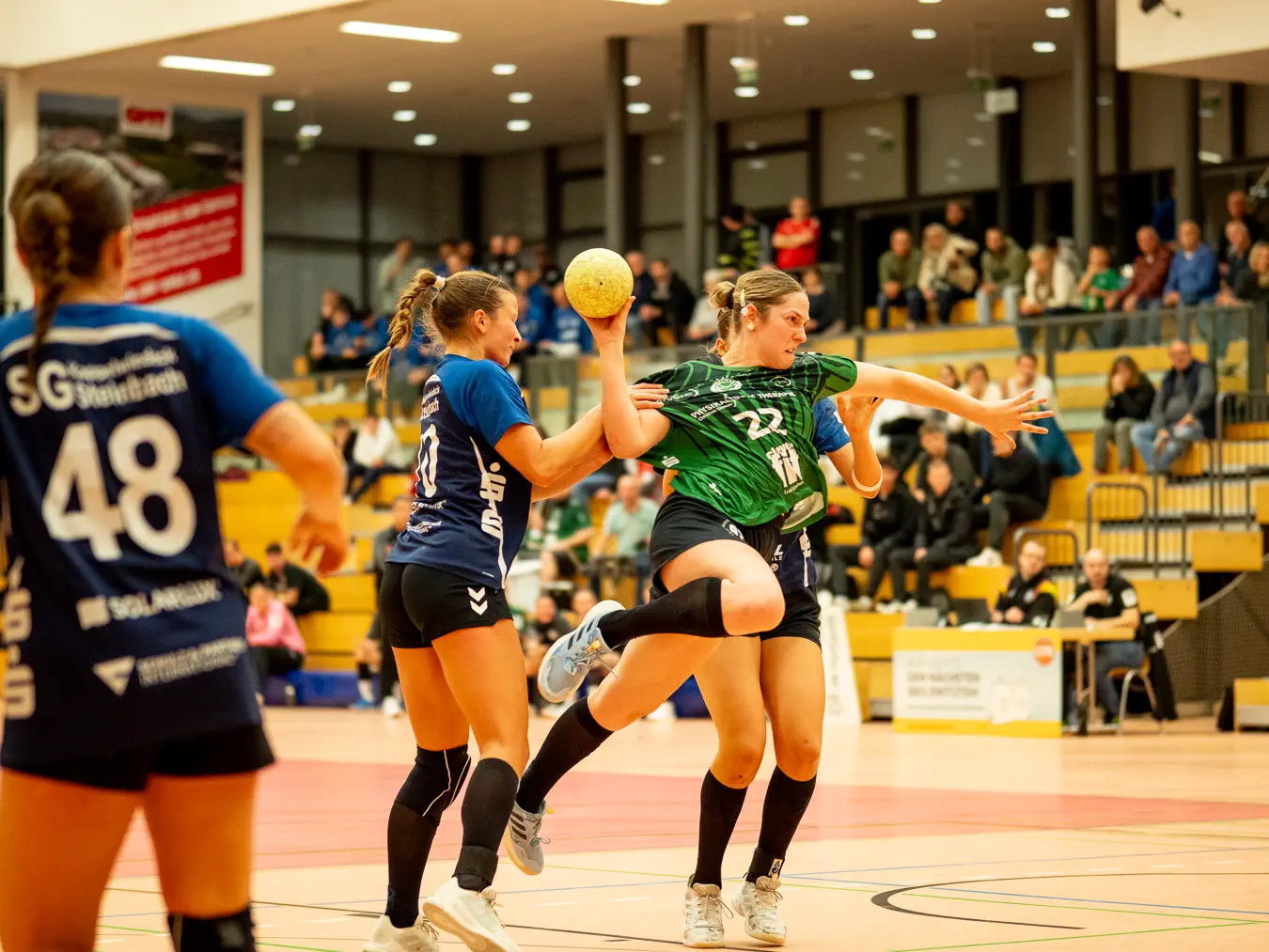 Eine Angreiferin mit Ball in der Hand im Sprung, zum Wurf ausholend. Eine Verteidigerin greift von hinten an die Schulter im Versuch, den Wurf zu verhindern.