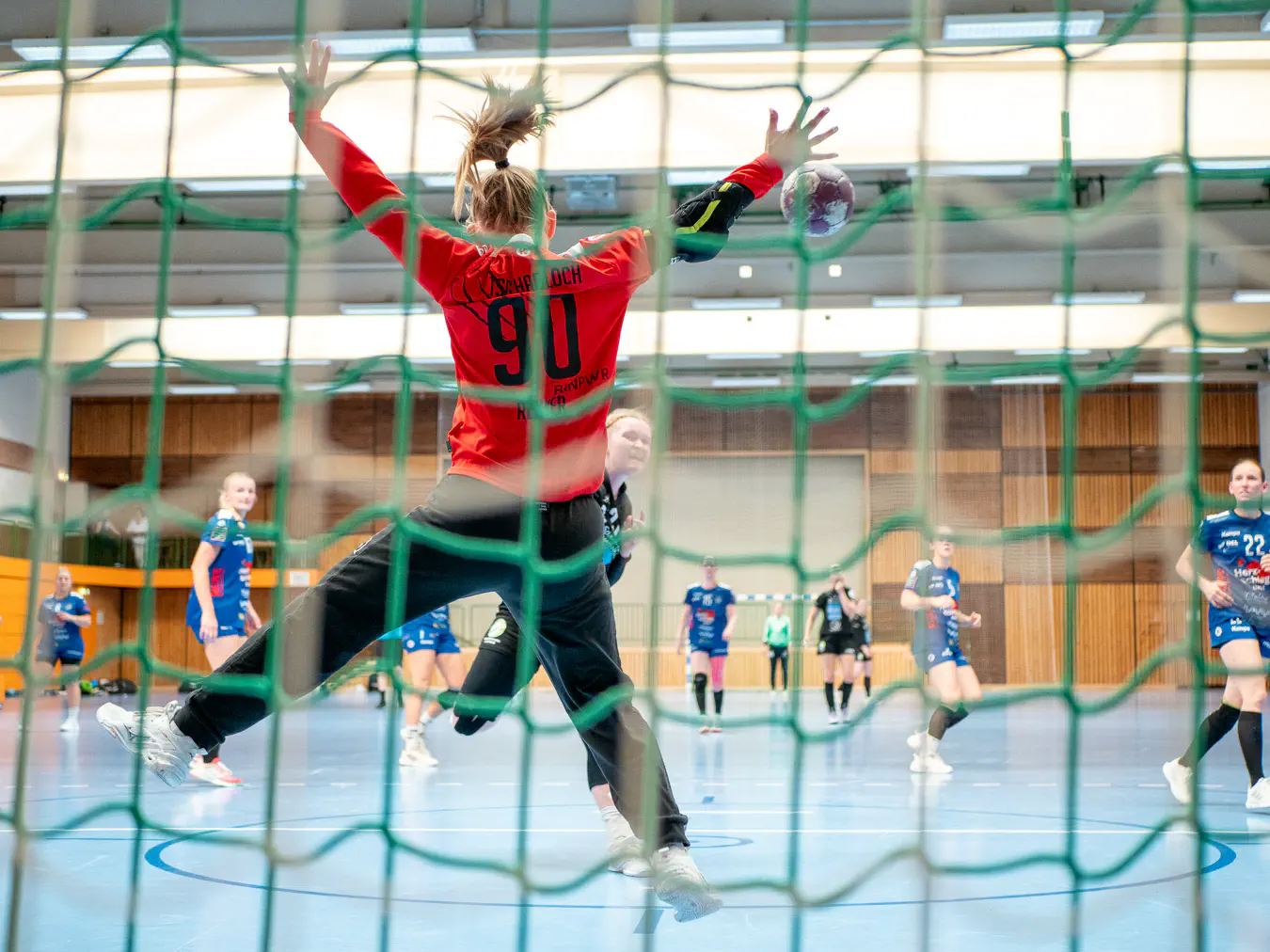 Eine Torfrau von hinten im Sprung, alle Viere von sich gestreckt, davor ein Ball im Flug auf sie zu, davor eine Angreiferin im Sprungwurf