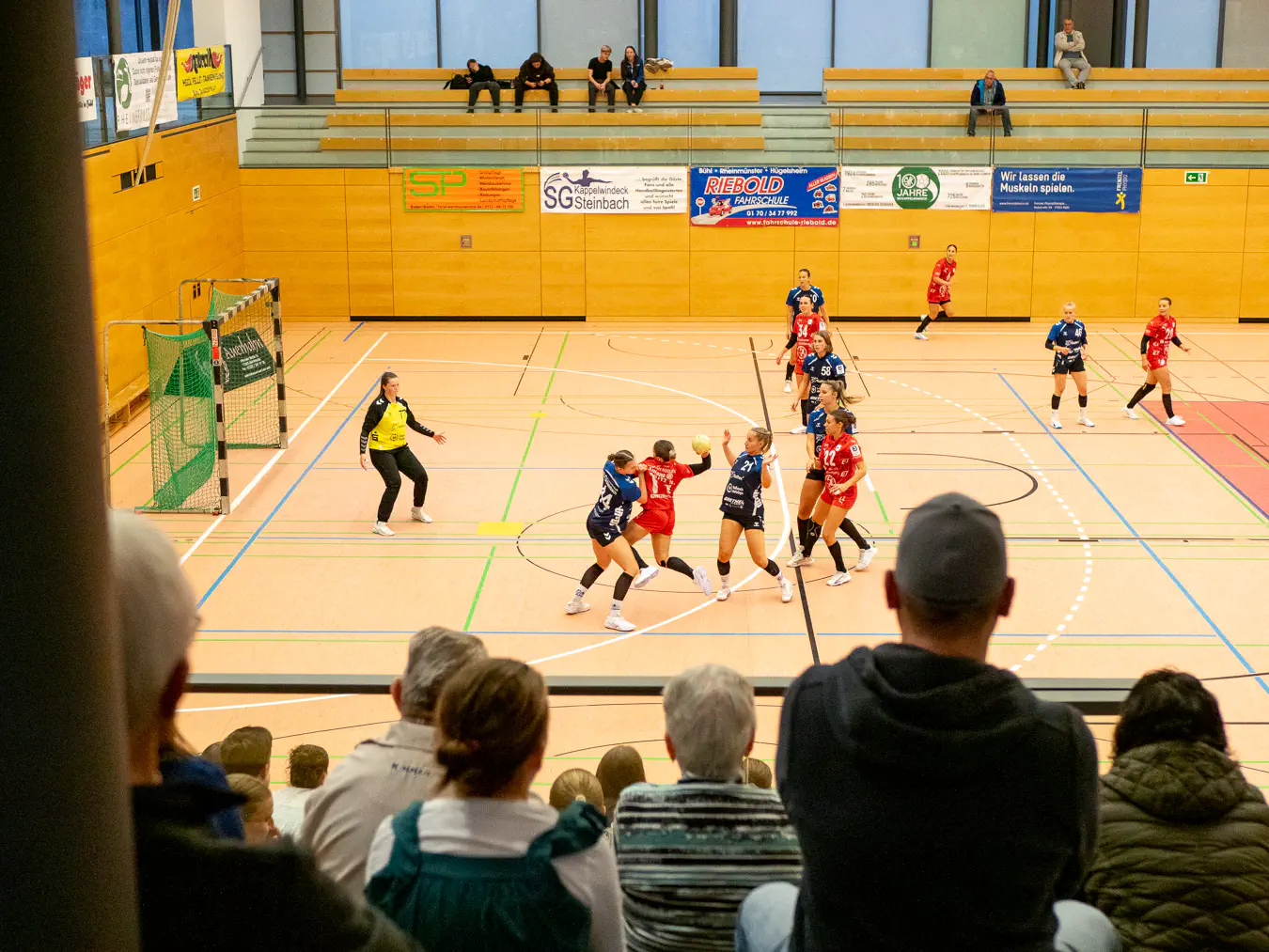 Menschen von hinten, die von einer Tribüne herab in eine Sporthalle schauen, wo eine Frau zwischen zwei anderen hindurch auf ein Handballtor zuspringt.